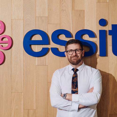 Essity employee stood infront of Essity logo background with arms folded and smiling Essity employee stood infront of Essity logo background with arms folded and smiling